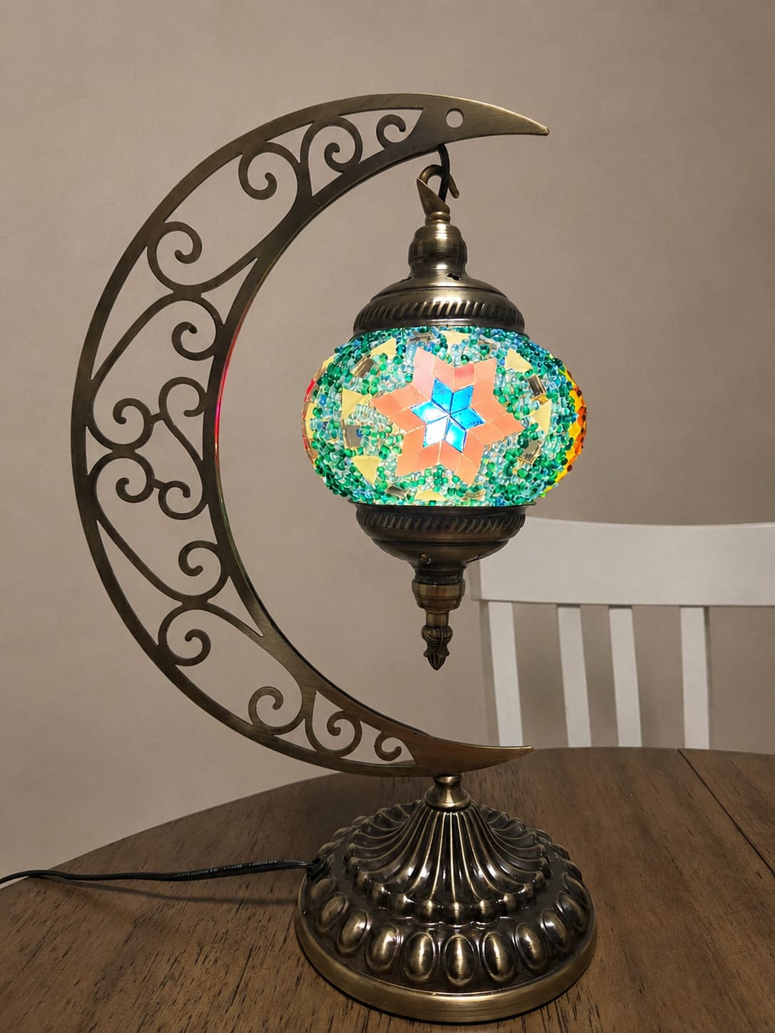 A decorative table lamp with a crescent moon frame in an antique bronze finish, holding a colorful mosaic glass globe shade with a star motif, set on a wooden table.