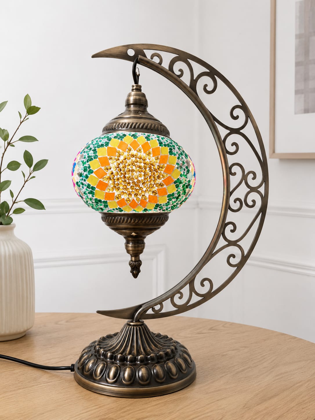 A decorative table lamp with a crescent-shaped ornate metal frame and a mosaic globe shade featuring a yellow sunflower design with green, blue, and orange details, displayed on a wooden table in a home setting.