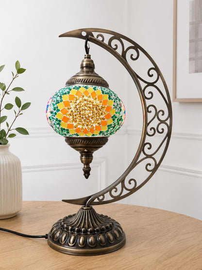 A decorative table lamp with a crescent-shaped ornate metal frame and a mosaic globe shade featuring a yellow sunflower design with green, blue, and orange details, displayed on a wooden table in a home setting.