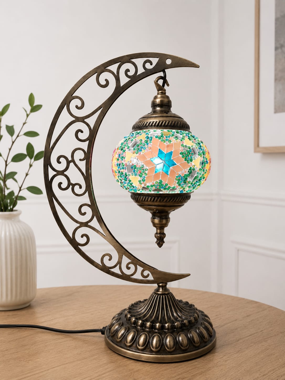 A decorative table lamp with an ornate crescent moon metal frame in an antique bronze finish, holding a colorful mosaic glass globe shade with a glowing star design, set on a wooden table in an indoor room.