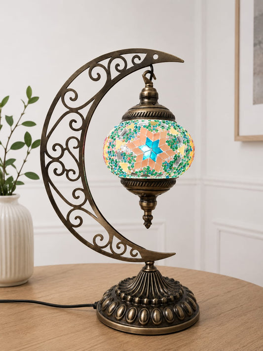 A decorative table lamp with an ornate crescent moon metal frame in an antique bronze finish, holding a colorful mosaic glass globe shade with a glowing star design, set on a wooden table in an indoor room.