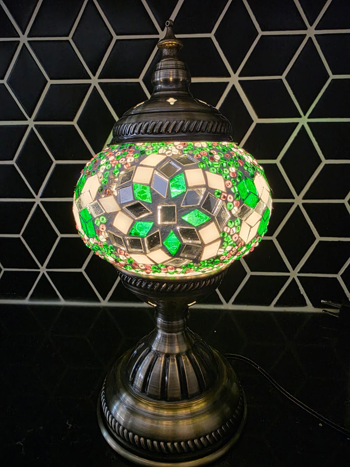 Decorative mosaic lamp with green and silver design against a geometric black and white tiled wall.